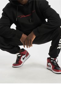 Felpa nera con cappuccio con scritta rossa "Rocawear", abbinata a pantaloni da jogging neri. Include sneaker Nike alte rosse e nere con dettagli bianchi.