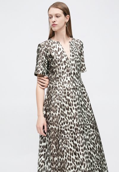 Γυναίκα που φοράει μακρύ φόρεμα με κοντά μανίκια, V-λαιμόκοψη και λεοπάρ μοτίβο, με σκουλαρίκια, όρθια μπροστά σε απλό λευκό φόντο.