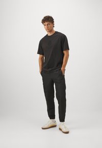 Svart bomullströja med en diskret runt logotyp på bröstet, i kombination med svarta cargobyxor och vita sportskor med bruna detaljer.