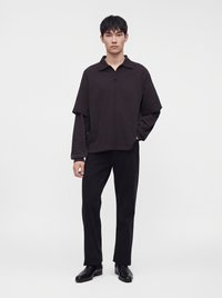 Homme asiatique mince debout portant un polo sombre à manches longues superposées, un pantalon noir et des chaussures en cuir noires sur un fond blanc uni.