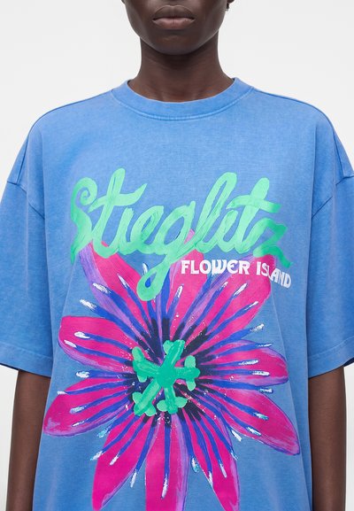 Άτομο με μπλε oversized T-shirt με το κείμενο «Stieglitz Flower Island» και μεγάλο ροζ και πράσινο λουλουδάτο σχέδιο στο μπροστινό μέρος.