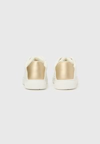Białe trampki z metalicznymi złotymi akcentami na pięcie. Górna część wykonana ze gładkiej skóry z teksturowaną podeszwą. Minimalistyczny design, zaokrąglony kształt.