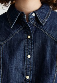 Denim shirt with a dark blue color, button-down front, pointed collar, and contrasting orange stitching detail. Features rounded white buttons.