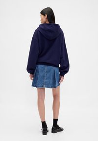Navy oversized hoodie met een capuchon met trekkoord, gecombineerd met een geplooide denim rok. Voorzien van robuuste mouwen en een losse pasvorm. Zwarte schoenen.