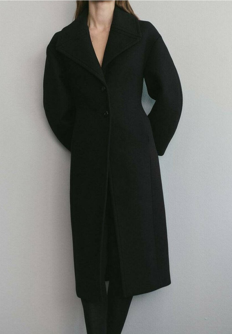 Femme portant un long manteau noir aux manches bouffantes exagérées, se tenant contre un mur gris clair uni.