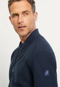 Dunkelblaues Bomberjackett mit strukturierter Oberfläche, Reißverschluss und geripptem Kragen. Besitzt ein kleines Logopatch am linken Ärmel.