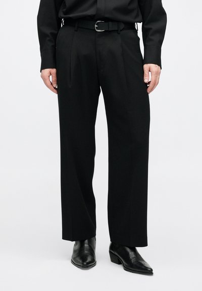 Μαύρο παντελόνι φόρμας με ραμμένη εφαρμογή, με πιέτες μπροστά, λεία υφή και συνοδευτική μαύρη ζώνη. Φορεμένο με μαύρες μπότες.