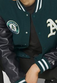 Veste universitaire verte en laine avec manches en cuir noir. Comprend le logo des Oakland Athletics et des lettres brodées blanches, des poignets rayés et des boutons-pression.