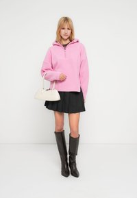 Ροζ φλις πουλόβερ με κουκούλα και φερμουάρ μέχρι τη μέση, σε συνδυασμό με μαύρη πλισέ φούστα και καφέ δερμάτινες μπότες μέχρι το γόνατο. Μπεζ τσάντα ως αξεσουάρ.