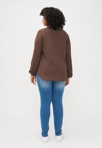 Une femme aux cheveux bouclés se tient de dos, portant un haut marron et un jean bleu, avec un fond neutre mettant en valeur sa tenue.
