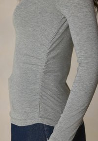 Graues Langarmshirt aus weichem Stoff mit seitlichen Raffungen. Verfügt über ein figurbetontes Design und einen Rundhalsausschnitt, kombiniert mit dunklem Denim.