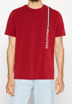 Mężczyzna w czerwonym, krótkim rękawie T-shircie z pionowym napisem „HILFIGER” i paskami po prawej stronie, w połączeniu z jasnoniebieskimi dżinsami.