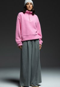 Übergroßer pinker Fleece-Pullover mit halbem Reißverschluss-Kragen, kombiniert mit grauen Streifen-Hose mit weitem Bein und einer hellgrauen Mütze.