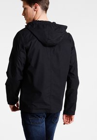 Black, hooded jacket with a smooth texture, featuring side seams and a slightly longer rear hem, paired with blue jeans.