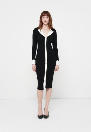 Robe noire en tricot avec un col blanc et un devant boutonné, présentant des manches longues et une silhouette ajustée, arrivant sous le genou.