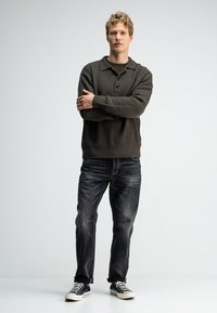 Dunkelgrüner Strickpullover mit gerippter Textur und dreiknöpfigem Verschluss, kombiniert mit ausgebleichten schwarzen Jeans und schwarzen Sneakers mit niedrigem Schaft.