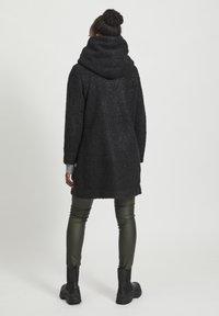 Svart, texturerad kappa med en stor huva, med en lös passform, sidofickor, och kombinerad med gröna, glänsande leggings och svarta stövlar.