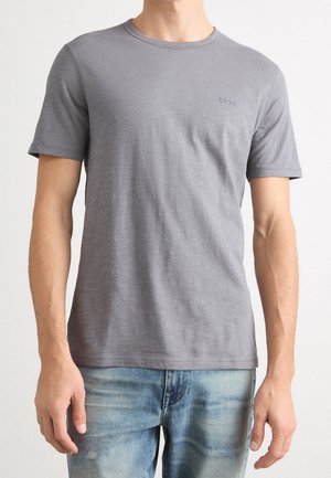 Mężczyzna ubrany w prostą szarą koszulkę z krótkim rękawem z subtelnym logo "BOSS" na piersi oraz wyblakłe niebieskie dżinsy, stojący na jednolitym tle.