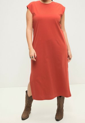 Robe rouge arrivant au genou avec des manches courtes, fabriquée en jersey doux ; dispose d'une coupe décontractée et de fentes sur les côtés. Portée avec des bottes de cow-boy marron.