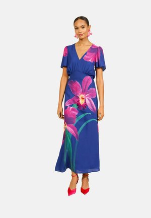 Kvinde iført en blå kjole med store pink blomsterprint, korte ærmer, V-hals og røde spidse hæle mod en ensfarvet baggrund.