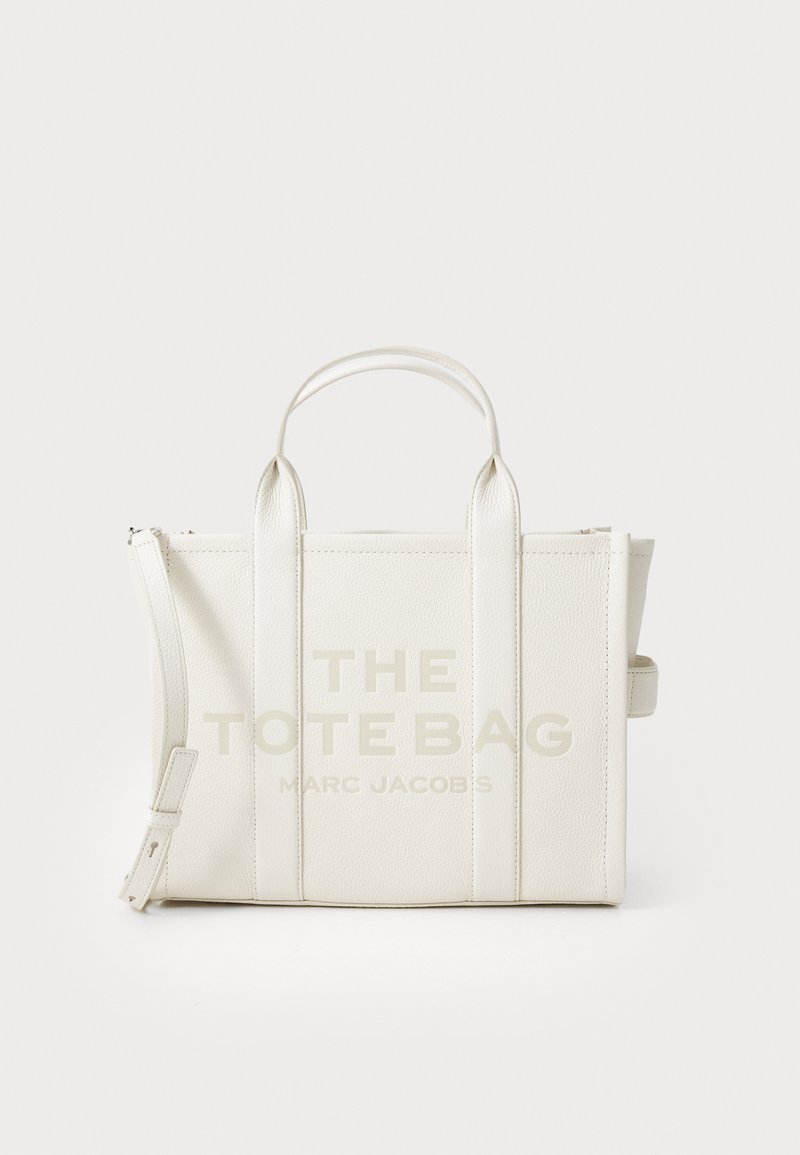 Geantă tote din piele albă, cu două mânere superioare, cu text imprimat "THE TOTE BAG MARC JACOBS" și închidere cu fermoar. Textură netedă, formă rectangulară.