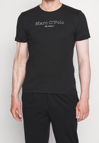 Hombre vestido con una camiseta negra de manga corta y pantalones negros, la camiseta tiene el texto "Marc O'Polo (Re-Nature)" en letras blancas.