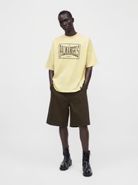 Άνδρας φορώντας κίτρινη oversized μπλούζα με κείμενο "Palm Angels", σκούρες καφέ βερμούδες μέχρι το γόνατο και μαύρα γυαλιστερά μποτάκια μέχρι τον αστράγαλο, στέκεται με τα χέρια στις τσέπες.