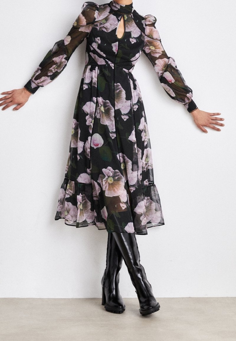 Robe florale noire avec de longues manches transparentes, un col en keyhole et un ourlet volé, ornée de fleurs roses et de feuilles vertes. Portée avec des bottes montantes.