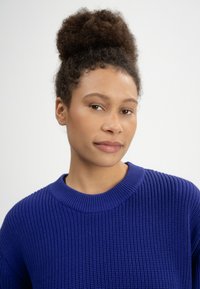 Blauer Strickpullover mit rundem Ausschnitt und strukturiertem Rippmuster. Nahaufnahme des Oberkörpers und Gesichts vor neutralem Hintergrund.