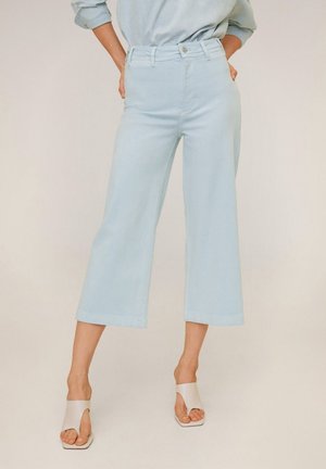 Persoon die lichtblauwe, wijde cropped jeans en witte, open teen sandalen met hak draagt tegen een neutrale achtergrond.