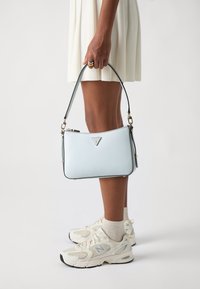 Sac à main en cuir bleu clair avec un logo argenté, de forme rectangulaire, et une fine bandoulière, porté par une personne vêtue d'une robe blanche et de baskets.