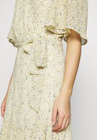 Květinové šaty v světle žluté barvě s volánovými rukávy. Mají páskový pas a jemné vzory v zelené a modré barvě. Měkká, splývavá textura.