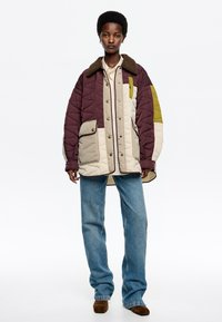 Chaqueta de plumas acolchada en burdeos, beige y verde oliva con grandes bolsillos frontales, un cuello de piel marrón y cierre de botones a presión.