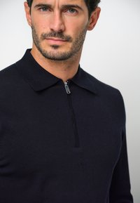 Marinblå zip-up polotröja, texturerad tyg, slim fit, framträdande krage, kort dragkedja. Enkel design med rena linjer och modern estetik.