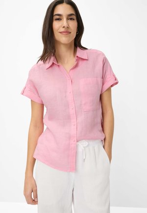 Frau trägt ein hellrosa kurzärmeliges Hemd mit Brusttasche und weiße Hose mit Kordelzug, steht vor einem weißen Hintergrund.