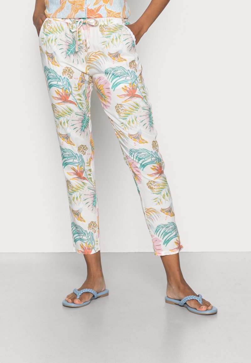 Femme portant un pantalon blanc avec des imprimés colorés de feuilles et de fleurs tropicales, assorti à des sandales plates tressées bleues.