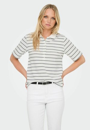 Polo bianca a maniche corte con righe blu navy, dotata di colletto e patta con tre bottoni. Indossata con pantaloni bianchi e una cintura nera.