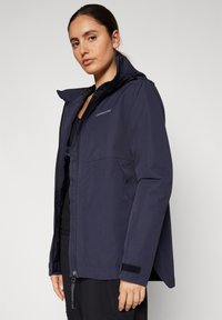 Granatowa wodoodporna kurtka z zamkiem błyskawicznym z przodu, regulowanym kapturem i mankietami na rzepy. Charakteryzuje się eleganckim designem oraz subtelnym logo na piersi.