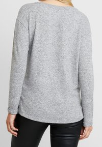 Femme portant un pull gris clair à manches longues et un pantalon en cuir noir, vue de dos avec ses cheveux blonds visibles.