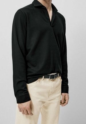 Homme portant un polo noir à manches longues rentré dans un pantalon beige avec une ceinture noire, sur un fond clair uni.