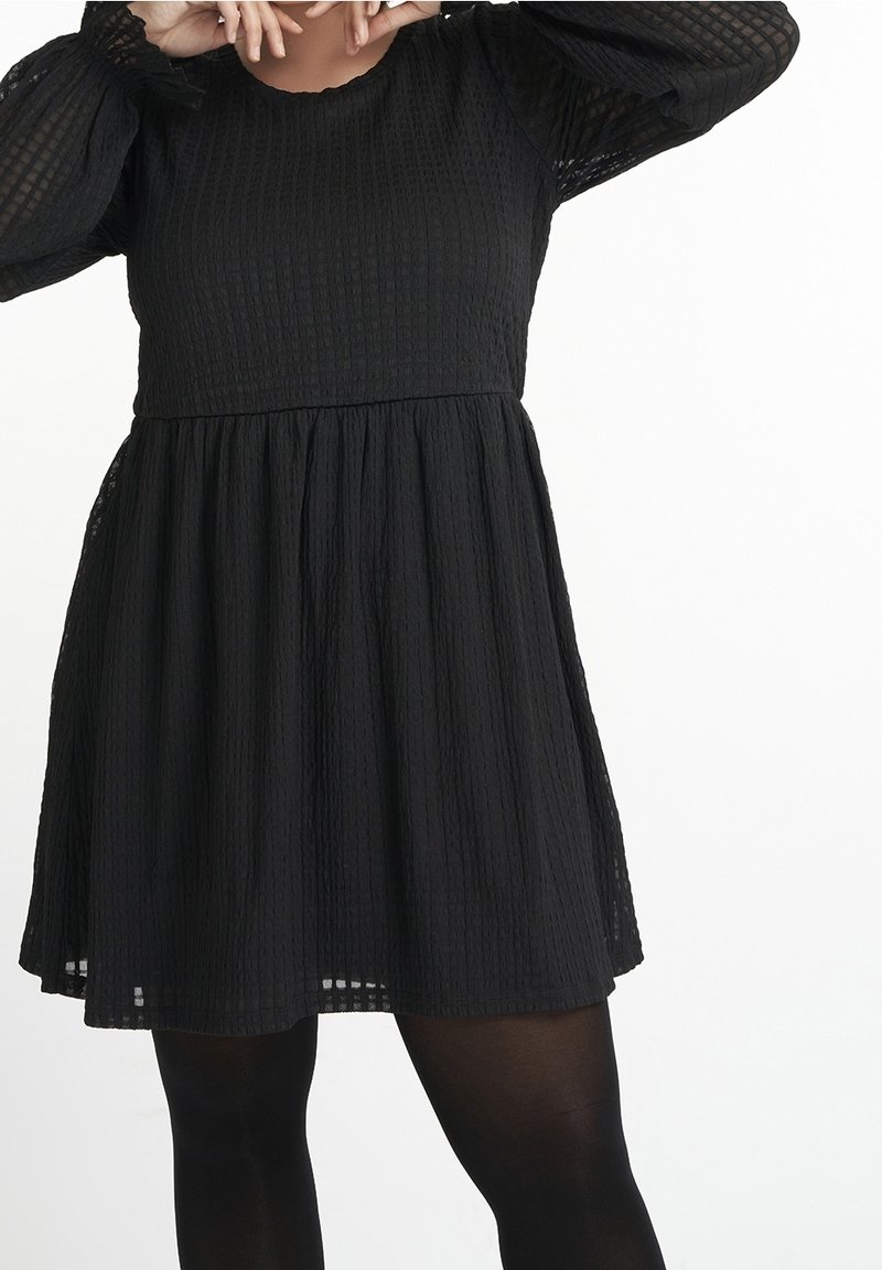 Femme portant une robe noire texturée à manches longues avec une jupe plissée et des collants noirs, posant avec les mains près du visage sur un fond blanc.