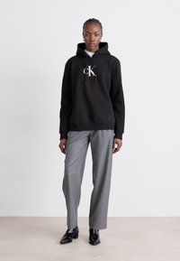 Felpa nera con cappuccio e grande logo bianco "CK", abbinata a pantaloni grigi su misura e scarpe nere a punta.