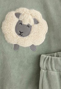 Grønt stof med en struktureret overflade har en flad cremefarvet fåre-applikation. Fåret har grå accenter og et simpelt smil. Inkluderer matchende bukser med omslåede ben.