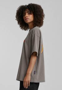 Camiseta gris de gran tamaño en algodón suave con mangas cortas, que presenta un logotipo naranja en el pecho y un dobladillo redondeado.