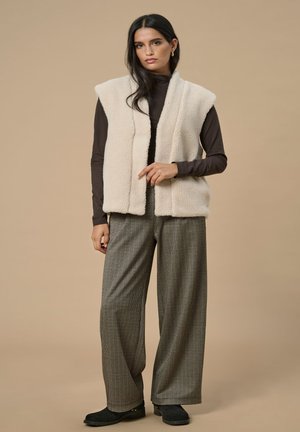 Crème neplederen gilets met een sjawl kraag, gecombineerd met een donkerbruine lange mouwen top en geruite wijde broekspijpen. Zwarte platte schoenen.
