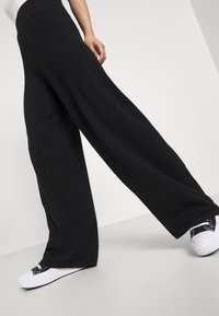 Black wide-leg pants made of soft fabric, feature a high waist and flowy design, paired with black and white sneakers.