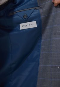Grau kariertes Sakko-Inneres mit blauer Fütterung, einem Knopf und einem "PIER•ONE"-Etikett. Enthält ausgeprägte Schneiderdetails.