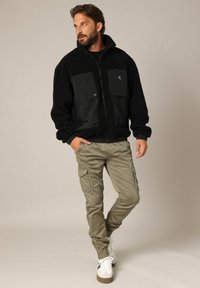 Zwarte fleecejack met twee grote zakken, gecombineerd met olijfgroene cargobroek. De outfit bevat witte sneakers met bruine accenten.