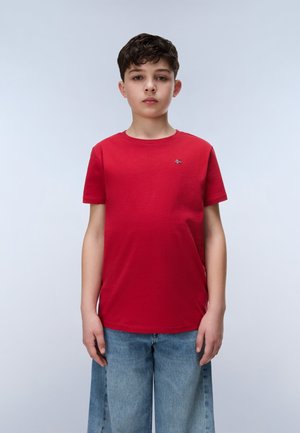 Garçon aux cheveux bruns courts portant un t-shirt rouge et un jean large bleu clair, debout devant un fond gris clair uni.