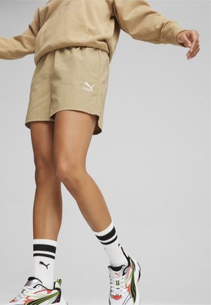 Shorts atletici bej, cu un brâu elastic și fante pe lateral, având un logo alb Puma. Purtați cu ciorapi albi și adidași colorați.